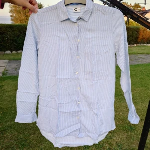 Blårandig skjorta från C, strl 34 - Snygg skjorta med tunna blå och vita ränder från C. Klassisk krage, bröstficka och knäppning framtill. Långärmad och tillverkad i ett lätt bomullstyg som känns fräscht och luftigt. Perfekt för en chill och stilren look.