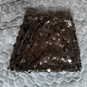 Svart paljettkjol med glittriga detaljer - Säljer en svart kortkjol täckt med stora glittriga paljetter som verkligen fångar ljuset. Kjolen har en rak och tight passform, perfekt för att sticka ut med en extra glam look. Materialet är syntet och paljetterna ger en cool och festlig vibe.