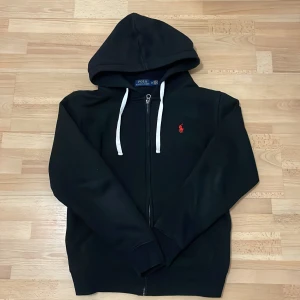Svart hoodie från Polo Ralph Lauren M - Svart hoodie från Polo Ralph Lauren med vit snörning i huvan och röd broderad logga på bröstet. Tröjan har dragkedja framtill och är långärmad. Klassisk och stilren modell som passar perfekt till jeans eller joggers. Priset kan diskuteras och förändras beroende på hur snabbt varan blir köpt.