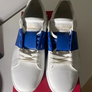 Valentino Garavani One Stud XL sneakers - Snygga Valentino Garavani One Stud XL sneakers i vitt skinn med en bred blå detalj över snörningen. Klassisk rund tå, platt sula och diskreta nitar baktill. Perfekta för dig som vill sticka ut med exklusiva och trendiga sneakers.