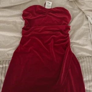 Röd sammetsklänning DK Harper L - Supersnygg röd sammetsklänning från DK Harper i kort modell. Axelbandslös design med lätt rynk vid bysten och figurnära passform. Perfekt för dig som vill sticka ut med en lyxig känsla och mjukt material.