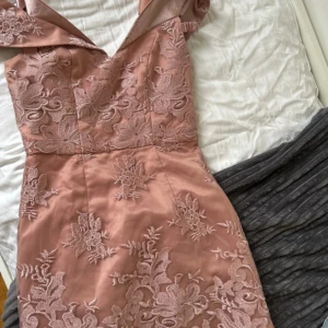 Rosa blommig långklänning med spets - Elegant långklänning i en mjuk rosa nyans med vackra blommiga spetsdetaljer. Klänningen har en v-ringad urringning och breda axelband. Perfekt för dig som vill sticka ut med en romantisk och sofistikerad look.