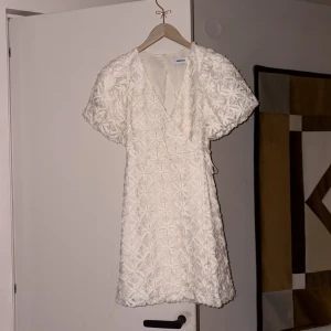 Vit blommig kortklänning Kappahl XS - Supersöt vit kortklänning från Kappahl i storlek XS. Klänningen har blommigt broderat tyg, omlott med v-ringning, puffiga kortärmar och en fin knyt-detalj i sidan. Öppen rygg ger en extra touch. Perfekt för sommarens alla tillfällen! Köpt för 950 kr och endast använd 1 gång vid konfirmation.