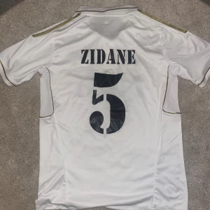 Real Madrid Zidane #5 Adidas tröja - Säljer en klassisk vit Real Madrid matchtröja från Adidas med Zidane och nummer 5 på ryggen. Tröjan har klubbmärke, Bwin-sponsor, guldiga detaljer och korta ärmar. Tillverkad i lätt och ventilerande ClimaCool-material. Perfekt för alla fotbollsfans! Size:S men ändå stor nog för att du som tar M också skulle kunna ta den. 