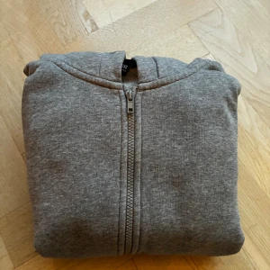Grå hoodie med dragkedja Only & Sons - Grå hoodie från Only & Sons med hel dragkedja framtill och huva. Tröjan har ribbade muddar, två fickor framtill och är tillverkad i mjuk bomullsblandning. Perfekt för chill dagar eller när du vill ha en enkel och stilren look.
