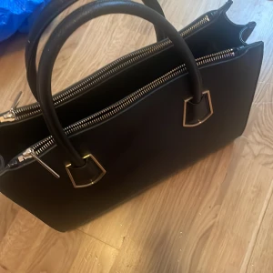 Svart handväska med gulddetaljer - Stilren svart handväska i skinnimitation med två rejäla handtag och tre stora fack med dragkedjor. Väskan har guldfärgade metalldetaljer och skyddande nitar undertill. Perfekt för dig som vill ha plats för allt och ändå se chic ut.