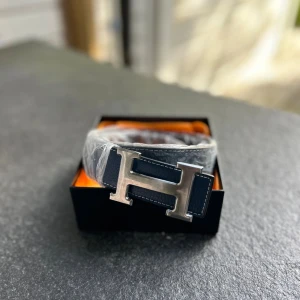 Bälte Mörkblå - Stilrent svart bälte från H i äkta skinn med ikoniskt H-formad metallspänne i silver. Bältet har vit kontrastsöm längs kanterna och levereras i originalask. Perfekt accessoar för att lyfta din outfit med en lyxig touch.