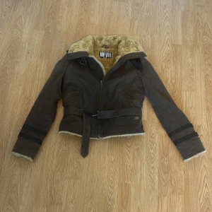 Brun skinnjacka med fuskpäls från Jofama - Snygg brun skinnjacka från Jofama med mjukt beige fuskpälsfoder, bälte i midjan och dragkedjor på fickorna. Jackan har markerad krage, spännen vid ärmslut och detaljer i mörkbrunt. Perfekt för kalla dagar och ger en cool look.