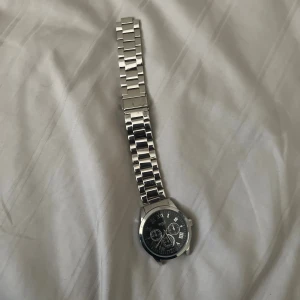 Silverfärgad regal klocka med länkarmband - Stilren klocka från regal med silverfärgat länkarmband i metall och svart urtavla. Urtavlan har romerska siffror och flera små visare för extra funktioner. Perfekt accessoar för dig som gillar klassisk och modern stil.