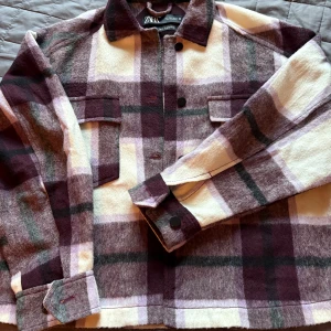 Rutig overshirt från Zara i ullmix - Snygg rutig overshirt från Zara i mjuk ullblandning. Jackan har stora bröstfickor med lock, svarta knappar och klassisk krage. Färgskalan går i vinrött, lila, beige och inslag av grått. Perfekt lager-på-lager-plagg för kyligare dagar.