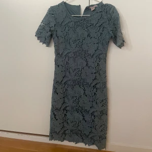 Blå spetsklänning med blommönster - Supersnygg blå klänning från Chi Chi London med blommig spets över hela klänningen. Modellen är knälång med korta ärmar och rund halsringning. Perfekt för dig som gillar romantiska detaljer och vill sticka ut lite extra.