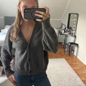 Mörkgrå kofta från Uniqlo - Mysig mörkgrå kofta från Uniqlo i storlek M. Koftan har v-ringning, stora knappar framtill och är tillverkad i ett stickat material som känns både mjukt och varmt. Perfekt för lager-på-lager och chill dagar. Långärmad och med avslappnad passform.