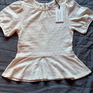 Vit blus med volang från Holly & Whyte - Supersöt vit blus från Holly & Whyte med korta puffärmar, volang nertill och broderade detaljer. Blusen har rund hals med virkad kant och knapp i nacken. Perfekt för dig som gillar romantisk och feminin stil.