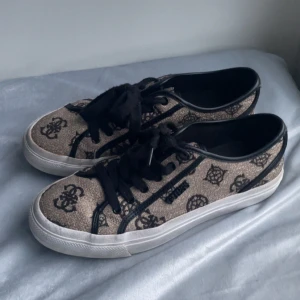 Beige och svarta sneakers från Guess - Snygga sneakers från Guess i beige tyg med svarta logomönster och svarta detaljer. Skorna har svarta snören, vit platt sula och rund tå. 