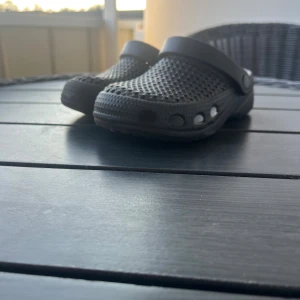 Svarta sandaler i gummi, strl 27/28 - Svarta sandaler i klassisk clog-modell med justerbar hälrem. Skorna är tillverkade i syntetmaterial och har ventilerande hål på ovandelen samt mönstrad sula för extra grepp. Perfekta för sommardagar eller som inneskor.