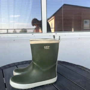 Gröna gummistövlar från 157 - Mörkgröna gummistövlar med vit kant och sula från 157. Klassisk modell med rund tå och dragflikar upptill för enkel påtagning. Perfekta för regniga dagar och festivaler. Snygg och enkel design som funkar till det mesta.
