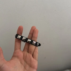 Svart valentino armband - Säljer ett smalt svart armbandet i skinn med silvriga pyramidformade nitar längs hela bandet. Armbandet har klassiskt spänne i metall och ger en edgy vibe till din outfit. Perfekt accessoar för att lyfta din stil med lite extra attityd. Helt ny bara använt ett par gånger kvitto och låda har tappats bort vid flytt