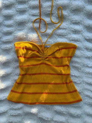 Randig orange halterneck topp Zara - Gul/orange ribbad halterneck topp från Zara med tunna knytband i nacken. Toppen har ett randigt mönster och är figurnära med lätt utsvängd nederkant. Perfekt för dig som vill ha en färgglad och trendig look i sommar. Ej använd storlek M