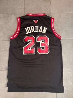 Chicago Bulls Jordan basketlinne - Snyggt svart basketlinne från 90 talet, Nike med Chicago Bulls-tryck och nummer 23 på både fram- och baksida. Röd och vit kant vid ärmar och hals, samt Jordan-tryck på ryggen. Perfekt för basket eller streetwear.
