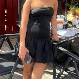 Svart kort of shoulder klänning - Snygg svart axelbandslös klänning med tight överdel och voluminös kjol. Kort modell som ger en trendig och ungdomlig look, perfekt för sommarens alla tillfällen. Enkel och stilren design som är lätt att matcha med accessoarer.