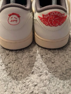 Nike Air Jordan 1 Low Travis Scott - Säljer ett par Nike Air Jordan 1 Low Travis Scott med ikonisk omvänd Swoosh i beige, grå mockadetaljer och vita läderpaneler. Baksidan har röda broderade loggor, inklusive Air Jordan Wings och Travis Scott smiley. Skorna har beige sula och vita skosnören.
