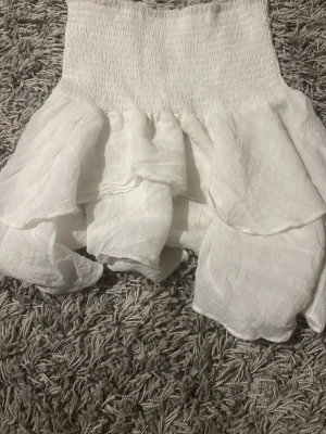 Vit volangkjol  - Supersöt vit kortkjol med flera lager volanger. Kjolen är gjord i ett lätt och luftigt tyg, perfekt för varma dagar. Passar perfekt till en avslappnad eller trendig look.