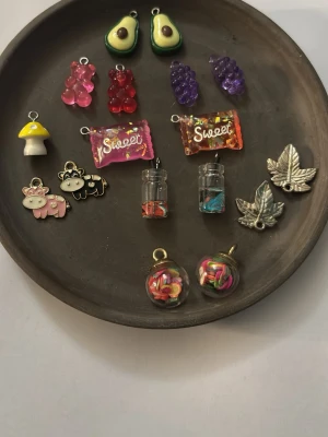 Mixade smyckesberlocker i olika former - Ett set med färgglada smyckesberlocker i form av avokado, godis, svamp, kossor, löv, vindruvor och små burkar. Materialen är blandade: metall, plast och glas. Perfekta för att skapa egna smycken med unik och lekfull stil.