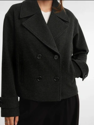 Mörkgrå kort kappa från Kappahl  - Kort kappa i mörkgrå ullmix från Kappahl. Ja Dubbelknäppt framtill med stora svarta knappar och bred krage. Ärmarna har dekorativa knappar och uppvik.  Köpt för 899kr 