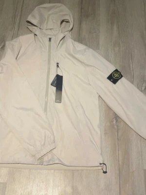 Beige vindjacka från Stone Island - Snygg beige vindjacka från Stone Island med huva och dragkedja framtill. Jackan har en bröstficka med dragkedja och den klassiska Stone Island-loggan på ärmen. Tillverkad i lätt och vindtätt material, perfekt för blåsiga dagar.