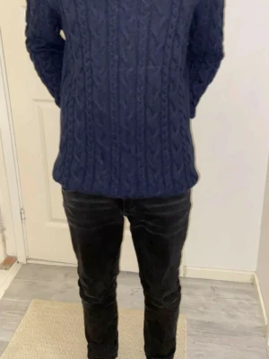 Mörkblå kabelstickad tröja - Mörkblå kabelstickad tröja med klassiskt flätmönster. Tröjan har rund halsringning och långa ärmar. Perfekt för kyliga dagar och passar till både jeans och chinos. Materialet ser ut att vara mjukt och värmande ullblandning.