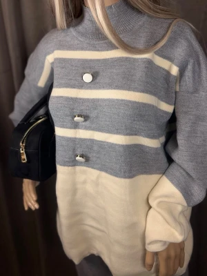 Randig stickad tröja med knappar - Mysig stickad tröja i ljusgrått och offwhite med breda ränder och tre dekorativa knappar framtill. Tröjan har hög hals och långa ärmar, perfekt för kyliga dagar. Materialet ser ut att vara mjukt och bekvämt, passar till många olika stilar.