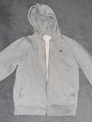 Grå hoodie från Lacoste - Snygg grå zip-hoodie från Lacoste med klassisk krokodillogga på bröstet. Tröjan har huva, dragkedja framtill och två fickor. Tillverkad i mjukt sweatshirtmaterial och har ribbade muddar vid ärmslut och nederkant.