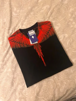 Svart t-shirt Marcelo Burlon med röd print - Svart t-shirt från Marcelo Burlon County of Milan med ikoniskt rött vingmönster över axlar och bröst. Rund halsringning och normal passform. Tillverkad i mjuk bomull, perfekt för en cool och edgy streetstyle-look.