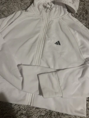 Vit hoodie med dragkedja från Adidas - Snygg vit hoodie från Adidas med dragkedja framtill och klassisk huva. Tillverkad i mjukt material som känns skönt mot huden. Diskret Adidas-logga på bröstet och ribbade muddar vid ärmsluten. Perfekt för en sportig och avslappnad stil.
