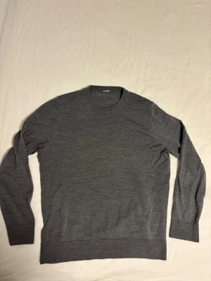 Arket merinoull-tröja - En stilren grå stickad tröja med rund hals och långa ärmar. Tillverkad i mjuk merinoull som känns skön mot huden. Tröjan har ribbade muddar vid ärmslut och nederkant för en klassisk look. Perfekt att matcha med jeans eller kjol.