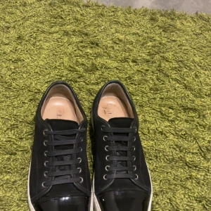 Lanvins  - Stilrena svarta sneakers från Lanvin med klassisk rund tå och svarta skosnören. Snörerna på ena skon har gått av men inte helt. Det sitter fortfarande bra och inget man märker. Skorna har en vit sula som kontrast och är tillverkade i skinn för en lyxig känsla. Perfekta för dig som gillar minimalistisk och tidlös design.