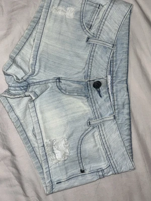 Ljusblå denimshorts med slitningar - Säljer ett par ljusblå vintage jeansshorts med slitna detaljer och klassisk femficksdesign. Shortsen har låg midja, bälteshällor och knappstängning framtill. Perfekta för varma dagar och en avslappnad stil. Säljer då de är förstora för mig! Säljer även dessa på vinted! Skriv till mig vid frågor!💕