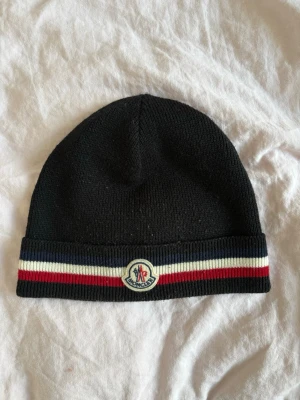 Svart Moncler mössa med ränder - Snygg svart mössa från Moncler med ribbad kant och klassiska Moncler-loggan framtill. Mössan har dekorativa ränder i blått, vitt och rött längs kanten. Tillverkad i mjukt stickat material som håller dig varm och stilren. Osäker om jag har kvittot kvar.