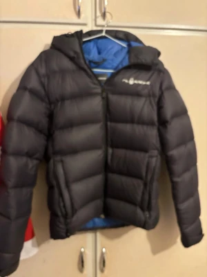 Svart dunjacka med huva från Sail Racing - Säljer en svart dunjacka från Sail Racing med huva och dragkedja framtill. Jackan har en puffig design, två sidofickor och logga på bröstet. Perfekt för kalla vinterdagar och riktigt skön att bära tack vare det mjuka fodret. Storlek xs men är stor i storlek, passar perfekt om man är runt 165-170cm. Inga större skador förutom lite av text borta på insidan och bokstaven S på bröstet.