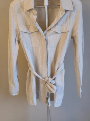 Beige skjorta med knytband - Stilren beige skjorta i mjukt tyg i linnemix med långa ärmar, klassisk krage och två bröstfickor. Skjortan har knappar framtill och ett matchande knytband i midjan för en snygg siluett. Perfekt för dig som gillar en enkel men trendig look.