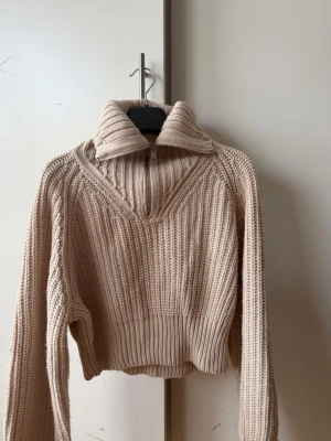 Beige stickad tröja med dragkedja - Säljer en beige stickad tröja med hög krage och dragkedja framtill. Tröjan har en croppad passform, bred ribbad mudd i nederkant och är tillverkad i ett tjockt, mjukt material. Perfekt för lager på lager och en avslappnad stil.