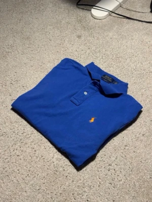 Blå pikétröja från Polo Ralph Lauren - Känns som en S skriv för mått.Klassisk blå pikétröja från Polo Ralph Lauren med gul broderad logga på bröstet. Tröjan har krage, knäppning med två knappar och är tillverkad i mjuk bomull. Perfekt till en avslappnad men ändå stilren look. Det är storlek M men känns som är S skriv för mått kan sitta som xs men den siter som S 