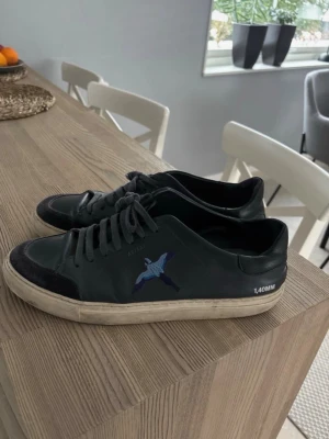  Axel Arigato skor - Snygga svarta sneakers från Axel Arigato med blå fågelbrodyr på sidan och vit sula. Skorna har svarta skosnören och detaljer i mocka på tån. Märkningen '140MM' syns på sidan. Perfekta för en clean och stilren look.