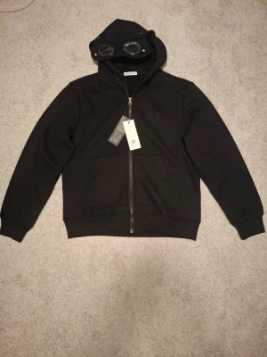 Svart hoodie från C.P. Company - Svart hoodie från C.P. Company med dragkedja framtill och klassiska goggles på huvan. Tillverkad i mjukt bomullsmaterial och har två stora fickor framtill. Diskret C.P. Company-logga broderad på bröstet. Perfekt för en streetwear-look.