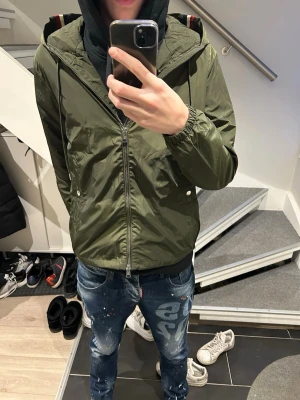 Olivgrön Moncler vindjacka med huva  - Snygg olivgrön vindjacka från Moncler med dragkedja, huva och elastiska muddar. Jackan har Moncler-logga på ärmen och röd-vita ränder på huvan. Tillverkad i lätt, glansigt material som passar perfekt till streetwear. Storlek 2