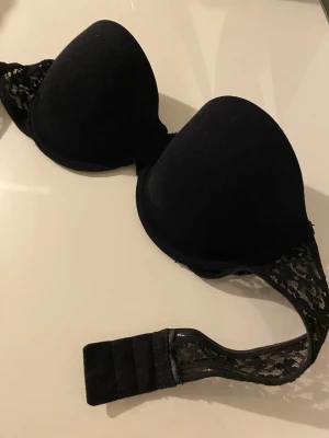 Svart push-up bh med spetsdetaljer - Snygg svart push-up bh med vadderade kupor och fina spetsdetaljer på sidorna och bandet. Bh:n har justerbar knäppning med tre hakar för bästa passform. Perfekt för dig som vill ha både komfort och stil.