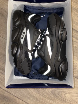 Svarta Dior B22 sneakers - Säljer ett par svarta Dior B22 sneakers med chunky sula och vita detaljer. Skorna har en futuristisk design med mesh och läderpaneler, samt reflekterande inslag. Klassisk snörning och logga på sidan. Perfekta för dig som gillar statement sneakers.