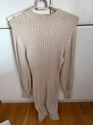 Beige ribbad långklänning - Säljer en beige ribbad långklänning med lång ärm och rund hals. Klänningen har en kroppsnära passform och är tillverkad i ett stickat material som ger en mjuk och bekväm känsla. Storlek M men passar även S