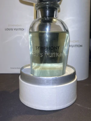 Louis Vuitton Symphony parfym ungefär 90 ml kvar - Exklusiv parfym från Louis Vuitton, Symphony. Flaskan är genomskinlig med rundad form och har en elegant silverfärgad metallkork med LV-logga. Parfymen har en ljusgul nyans och levereras i en stilren vit förpackning.