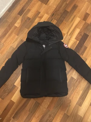 Svart pufferjacka från Canada Goose - Säljer en svart pufferjacka från Canada Goose med stor huva och klassisk logga på ärmen. Jackan har två stora fickor framtill, dragkedja och knappar samt ribbade muddar vid ärmsluten. Perfekt för kalla vinterdagar och riktigt varm. Storleken är L men den passar även som M. Först till kvarn! 